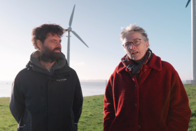 Twee mensen staan op een eiland vol met gras en twee windmolens op de achtergrond